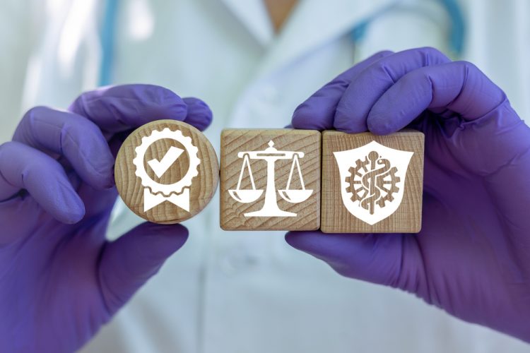 idea of pharmaceutical or clinical trial regulation: close up of a doctors gloved hands holding up three wooden blocks with symbols for regelation or legislation, medicine and approval or certification