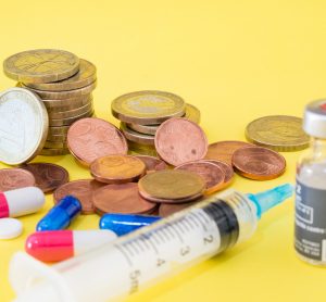 vial of vaccine with syringe and pills next to stacks of euro coins