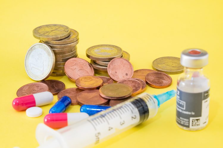 vial of vaccine with syringe and pills next to stacks of euro coins