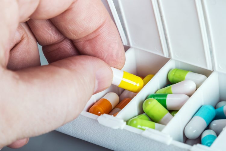 hand lifting yellow and white capsule from a sectioned drug box with other capsules of different colours in it