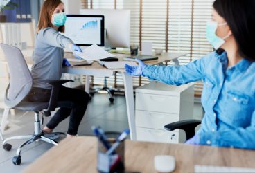 two members of staff wearing face masks and passing a sheet of paper while keeping their distance