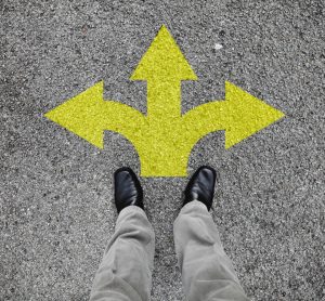 A pair of feet standing on a tarmac road with yellow arrow print pointing in three different directions - idea of decision making/guidance