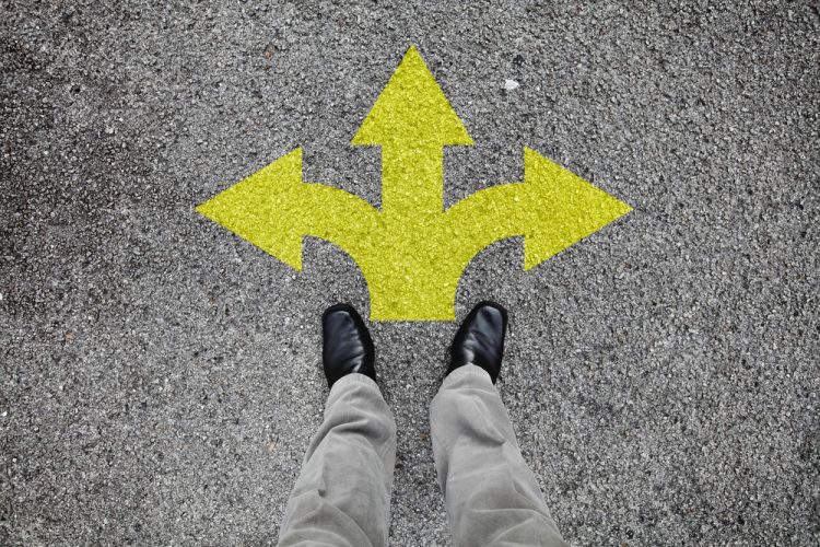 A pair of feet standing on a tarmac road with yellow arrow print pointing in three different directions - idea of decision making/guidance