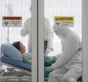 patient in a hospital bed with doctors in personal protective equipment behind glass doors with quarantine stickers on