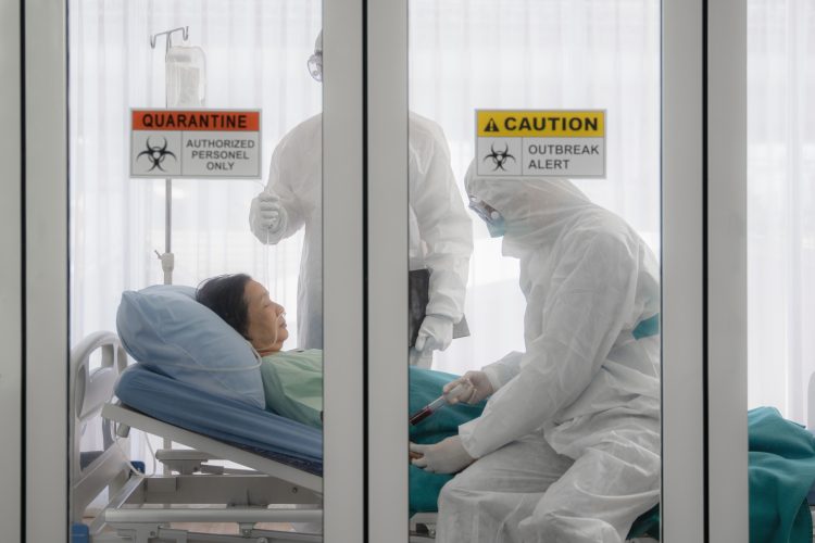 patient in a hospital bed with doctors in personal protective equipment behind glass doors with quarantine stickers on