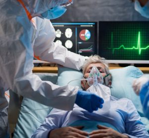 old man in a hospital bed on a ventilator surrounded by monitors and doctors wearing PPE