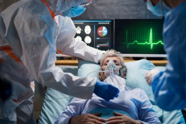 old man in a hospital bed on a ventilator surrounded by monitors and doctors wearing PPE