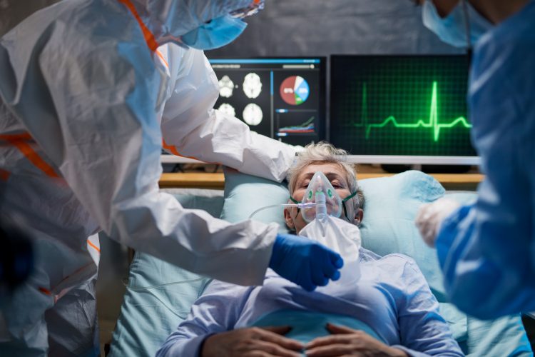 old man in a hospital bed on a ventilator surrounded by monitors and doctors wearing PPE