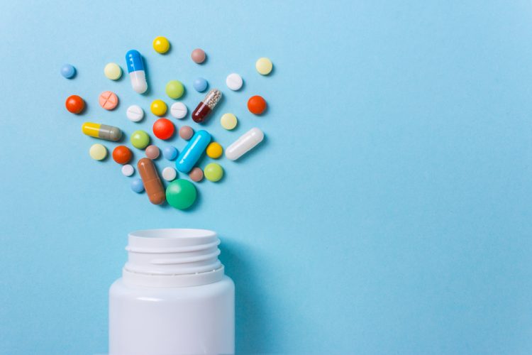 Assorted pharmaceutical pills, tablets and capsules spilling from a tipped over white bottle on a blue background