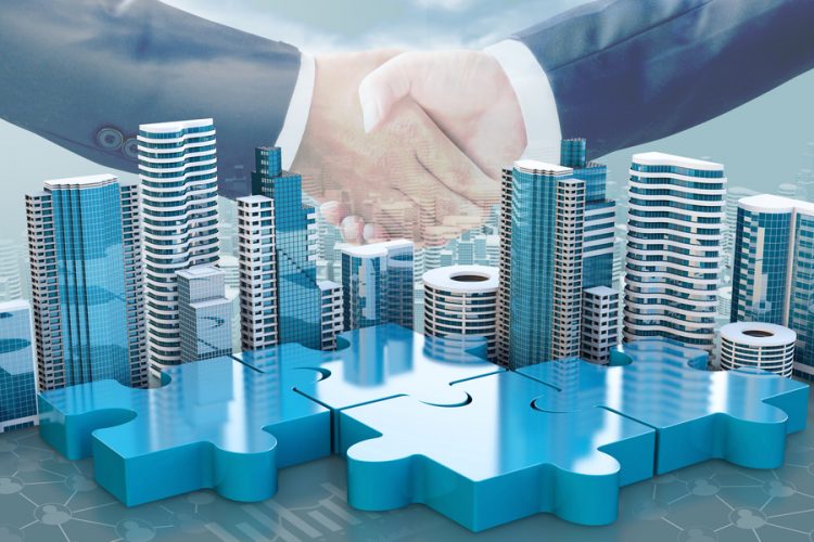 merger and acquisition business concepts - puzzle pieces with buildings on top linking together under two suited businesspeople shaking hands