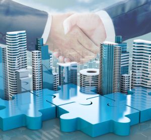 merger and acquisition business concepts - puzzle pieces with buildings on top linking together under two suited businesspeople shaking hands