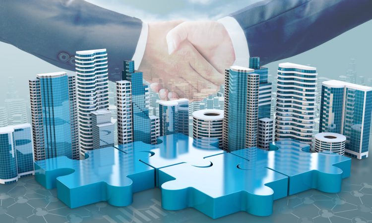 merger and acquisition business concepts - puzzle pieces with buildings on top linking together under two suited businesspeople shaking hands