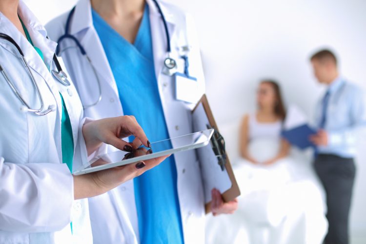 Young woman doctor holding a tablet and conferring with colleague with patient in bed blurred in background