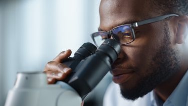 close up of a black male pharmaceutical microbiologist looking down a microscope - idea of microbial contamination