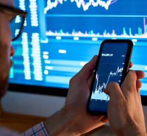 Man on phone looking at share prices on phone and computer screen