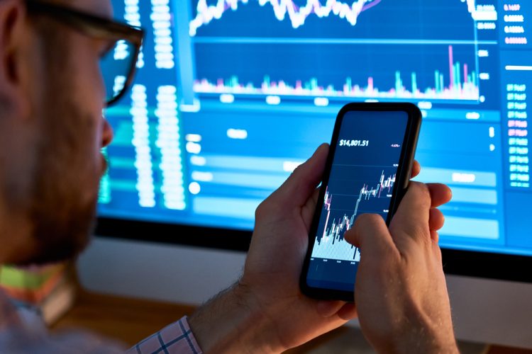 Man on phone looking at share prices on phone and computer screen