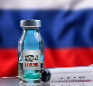 vial labelled 'COVID-19 Vaccine Sputnik' and syringe in front of the Russian Flag (white, blue red horizontal stripes)