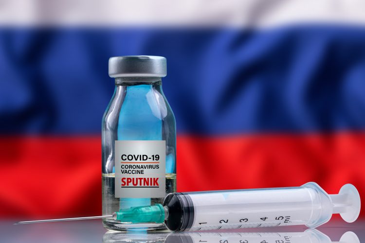 vial labelled 'COVID-19 Vaccine Sputnik' and syringe in front of the Russian Flag (white, blue red horizontal stripes)