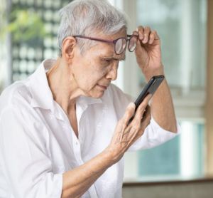 Elderly Asian woman lifting glasses as she struggles to read text on her phone - idea of vision loss in the elderly such as through age-related macular degeneration