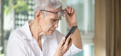 Elderly Asian woman lifting glasses as she struggles to read text on her phone - idea of vision loss in the elderly such as through age-related macular degeneration