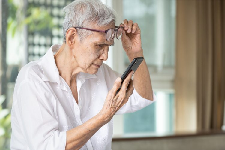 Elderly Asian woman lifting glasses as she struggles to read text on her phone - idea of vision loss in the elderly such as through age-related macular degeneration