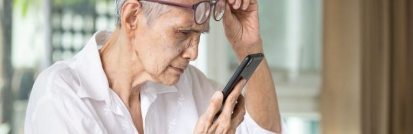 Elderly Asian woman lifting glasses as she struggles to read text on her phone - idea of vision loss in the elderly such as through age-related macular degeneration